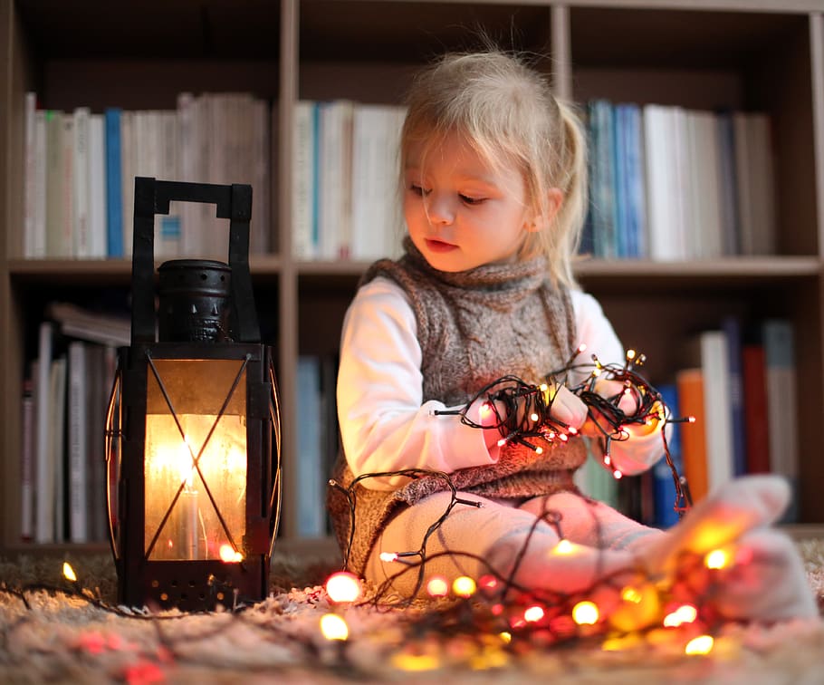girl in knit vest sits beside lighted lantern and holds turned on string lights