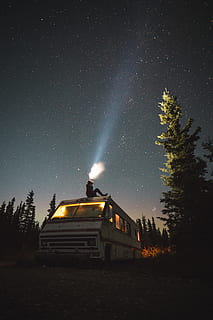 person sits on top of van at night, star, headlight, tree, camper HD wallpaper