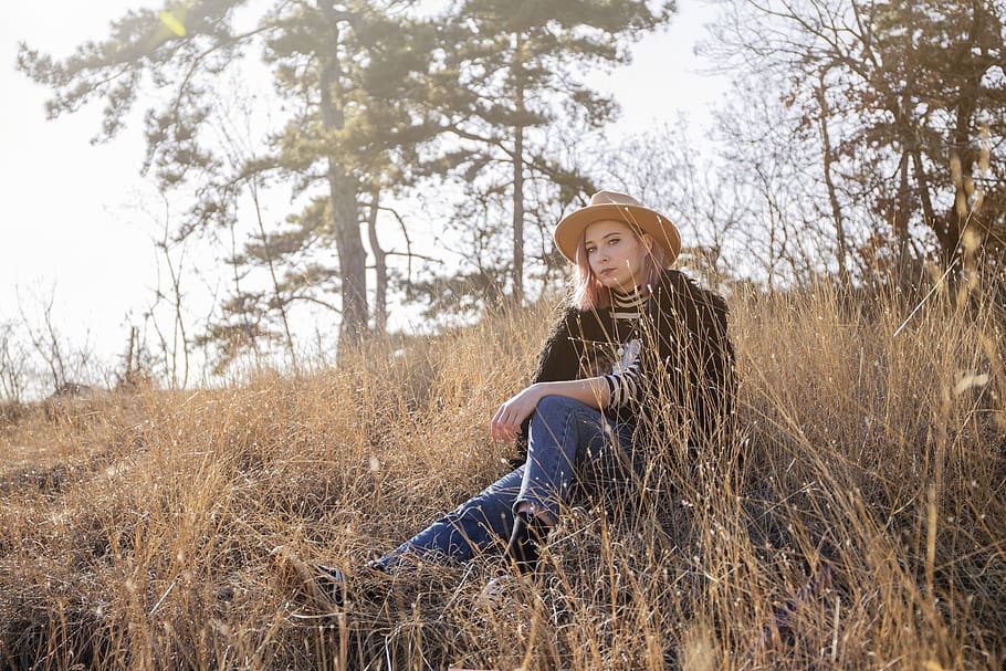 woman wearing brown sun hat, black jacket, and blue jeans sitting on slope of grass during daytime