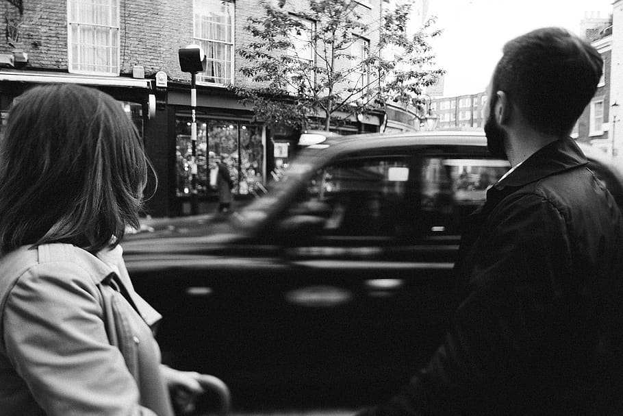 Grayscale Photography Man and Woman Standing While Cars on Road Near Buildings