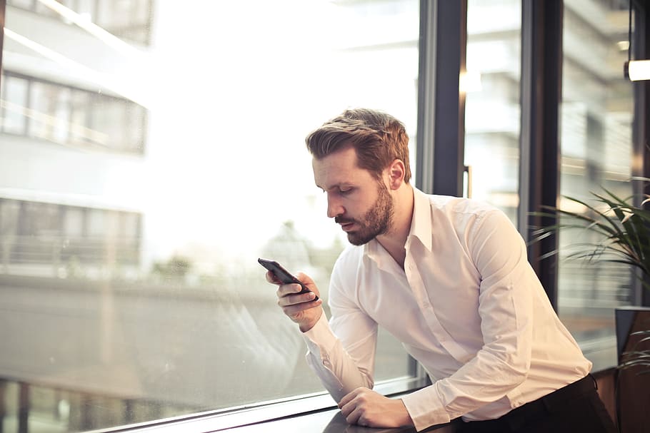 Young Adult Man in White Shirt Checking His Phone Near Window In Office