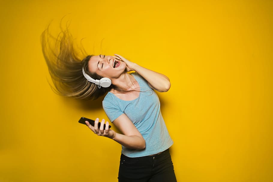 Excited woman dancing and listening music with headphones and smart phone with hair moving, isolated on yellow background