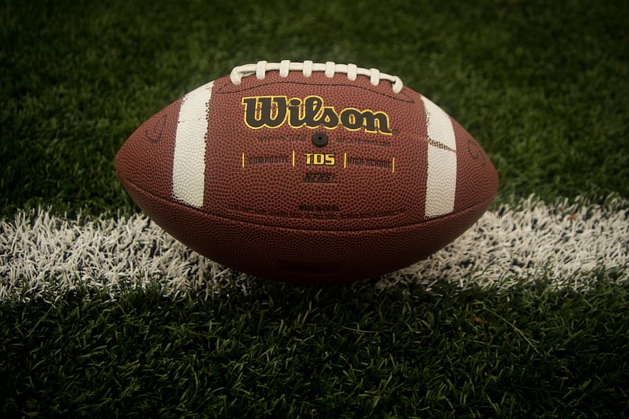 This picture shows an american football manufactured by Wilson lying on a football field. The football has the typical brown and redish color with some white lines painted on it and white laces on the top., HD wallpaper