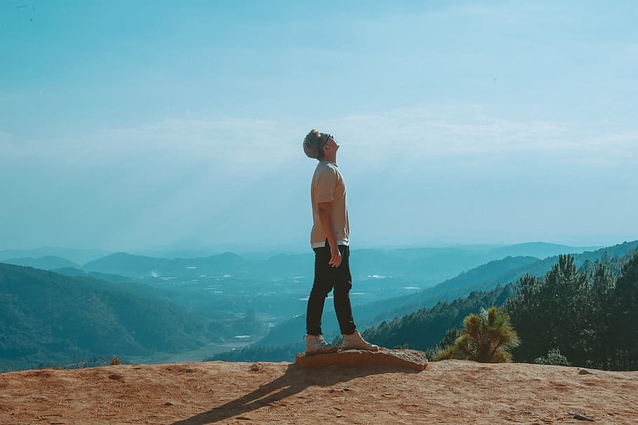 Man Wearing Beige Shirt and Black Pants Looking Up With Blue Sky and Mountains in the Background HD wallpaper