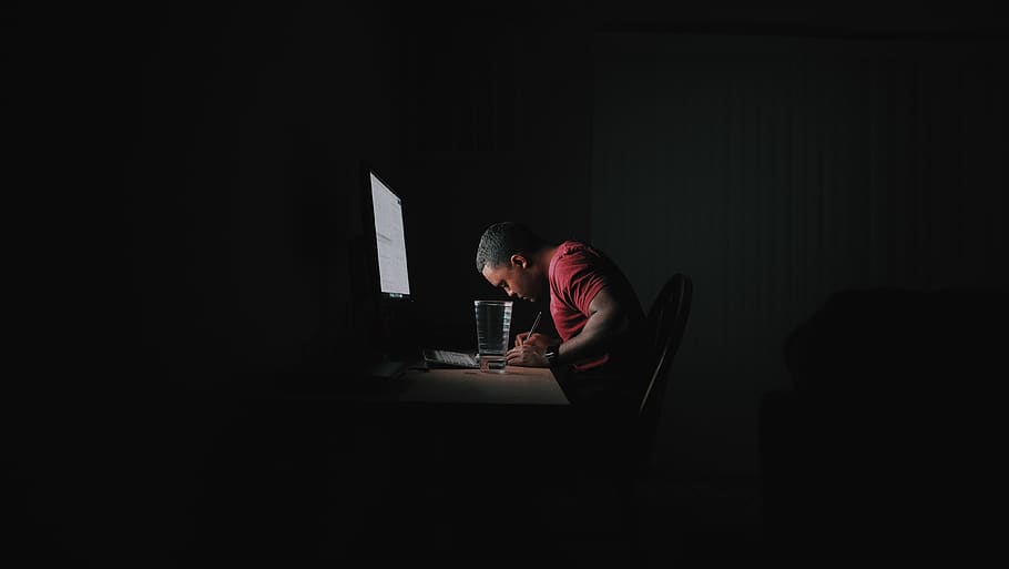 man in red shirt writing while leaning on desk in front of computer monitor