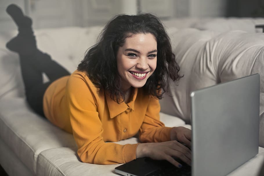 A young woman in yellow button-up shirt and black leggings smiling while working on her laptop on the white couch