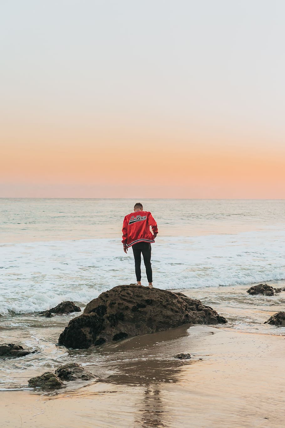 man wearing orange jacket standing on seashore rock near beach HD wallpaper