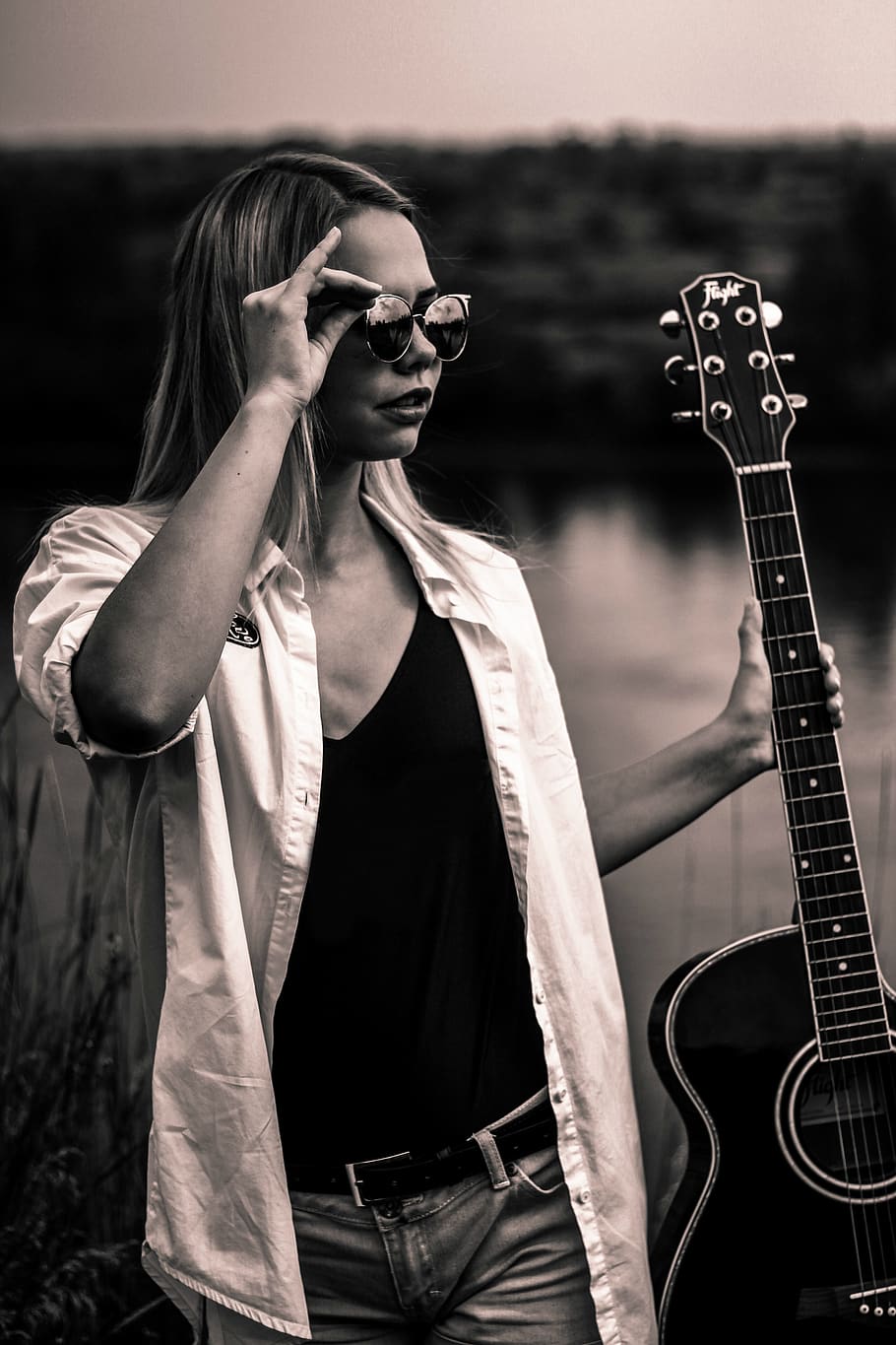 grayscale photo of woman wearing deep V-neck top and button-up long-sleeved shirt while holding sunglasses and acoustic guitar