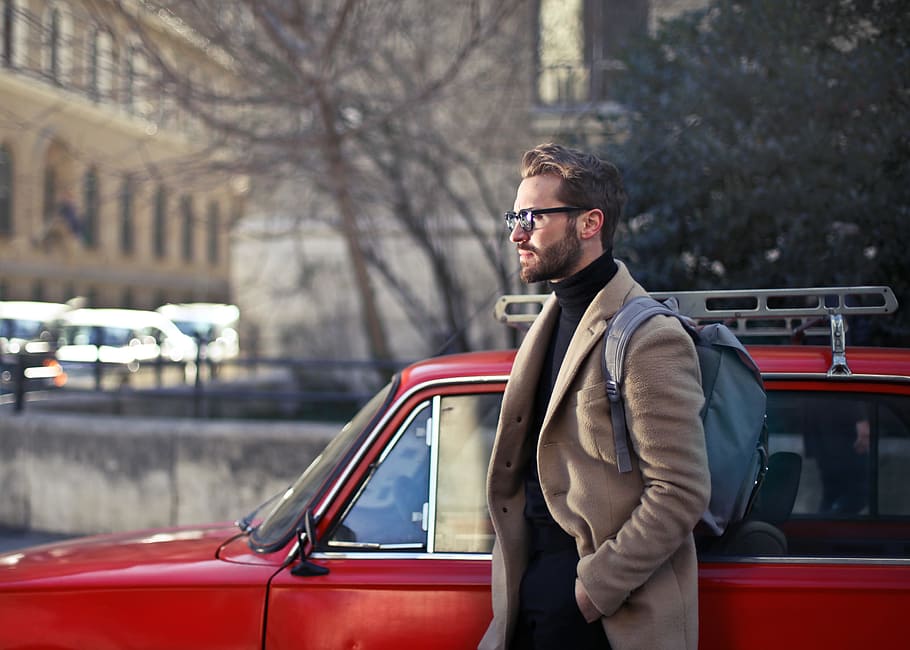 Young Adult Man in beige jacket with backpack on his shoulders,walking on the road as red vintage car seen beside him