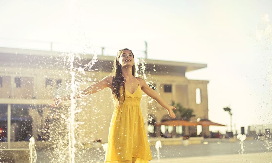 Beautiful young woman in yellow spaghetti strap dress dancing under the fountain on the street