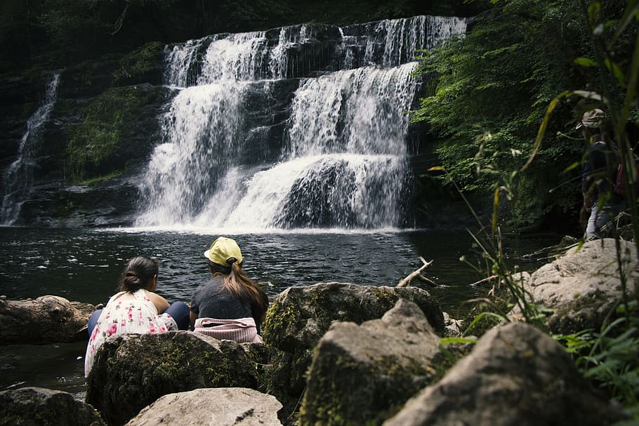 Two Women Sitting on Rock Near Waterfalls, boulders, cap, daytime HD wallpaper