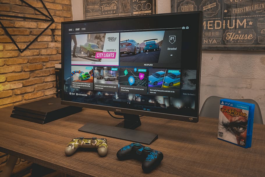 Image of a 4K monitor setup to play games on, with Need For Speed: Heat being shown on its screen and 2 PS4 controllers sat on the table in front