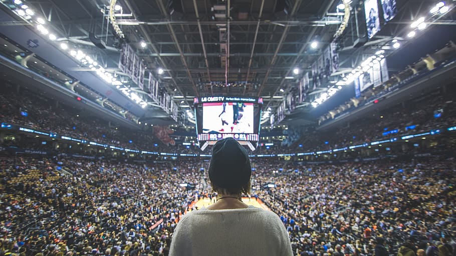 Back view of a fan wearing a black cap at a basketball game with crowds in the stands and a big LED screen in the background