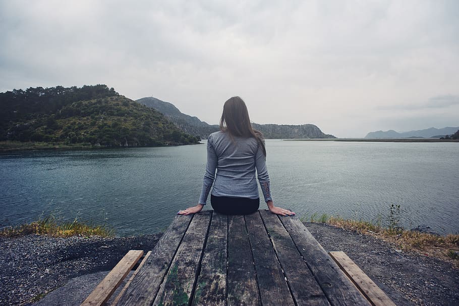 Woman Wearing Gray Long-sleeved Shirt and Black Black Bottoms Outfit Sitting on Gray Wooden Picnic Table Facing Towards Calm Body of Water at Daytime HD wallpaper