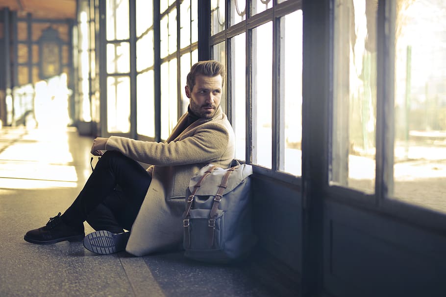 Man Wearing Brown Coat Beside White Backpack Sitting Near Window HD wallpaper