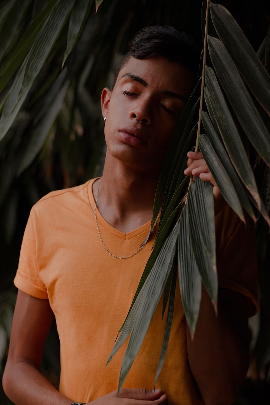 Photo of Boy in Orange V-neck T-shirt Posing Next To Green Leafed Plant With His Eyes Closed