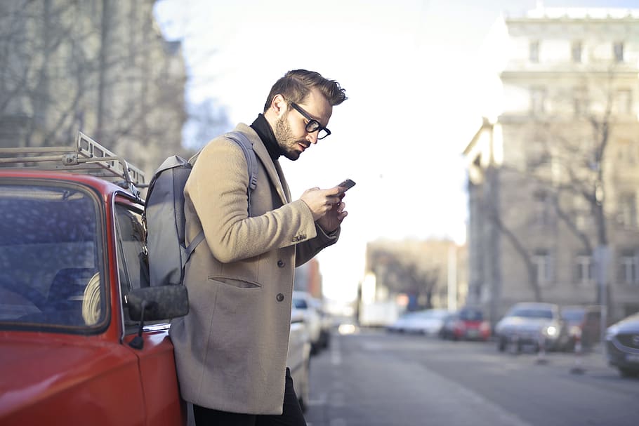 Man in Beige Coat Holding Phone Leaning on Red Vehicle, backpack, HD wallpaper