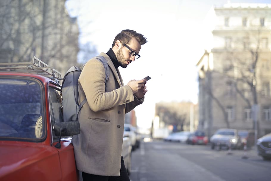 Man in Beige Coat Holding Phone Leaning on Red Vehicle, backpack HD wallpaper