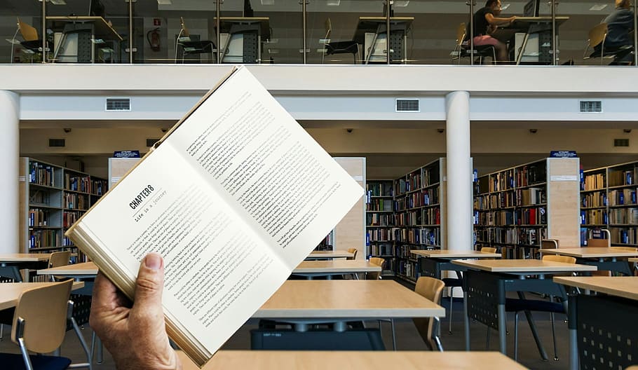 Photo illustration of book being held up in a library with empty desks HD wallpaper