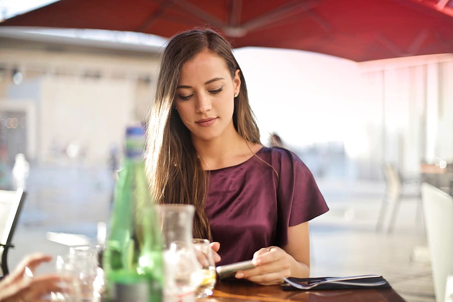A young blonde woman wearing a purple red dress looking at her mobile phone screen at a restaurant