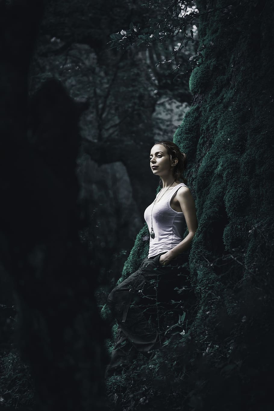 woman in white tank top stands while leaning on green moss covered tree trunk HD wallpaper