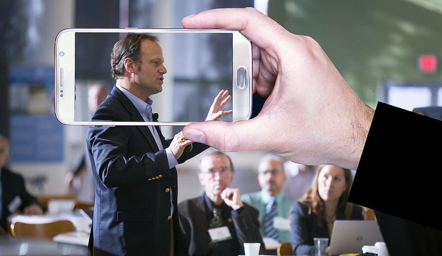 Photo illustration of taking a video or photo of a person speaking in public.