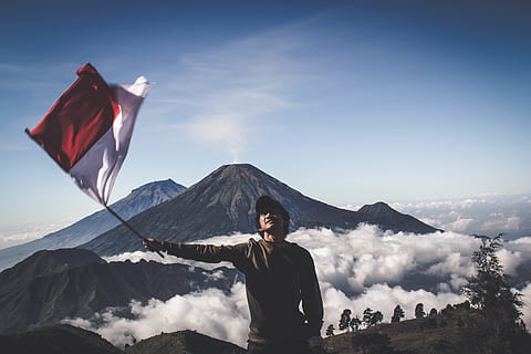 Man Wearing Black Crew-neck Sweater Holding White and Red Flag Standing Near Mountain Under Blue and White Sky HD wallpaper