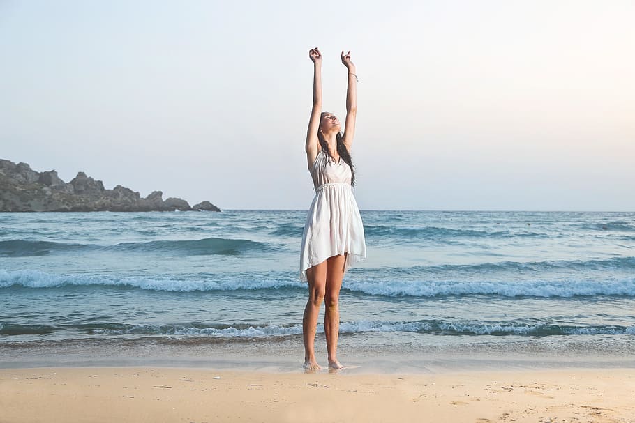 A young brunette woman wearing a white dress standing on the beach with her hands up