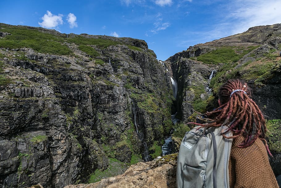 Person Wearing Brown Sweater and Grey Backpack With Brown Hair Dreadlocks on Top of Mountain Overlooking Waterfalls HD wallpaper