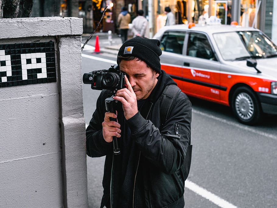 Man Holding Camera, beanie, bonnet, car, city, depth of field HD wallpaper