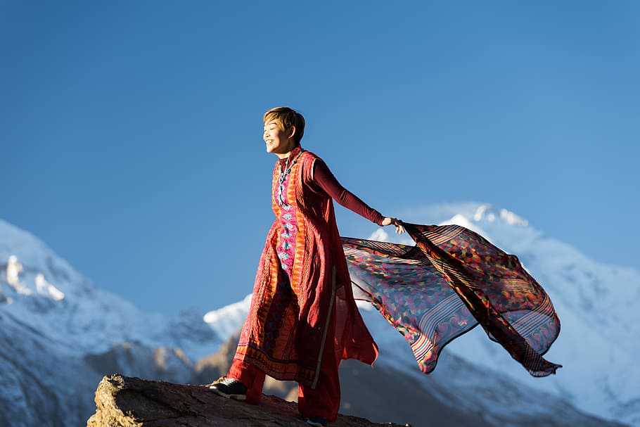 Woman in Orange Dress Standing on Top of Rock Cliff Holding Scarf Near Mountain Covered With Snow HD wallpaper