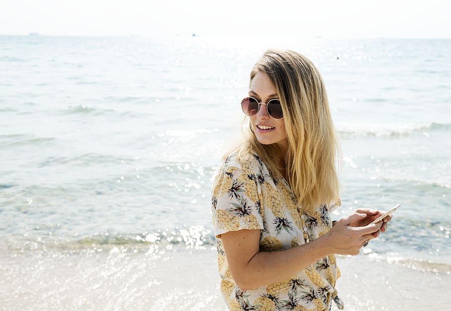 Woman in Black Yellow and White Floral Button-up Shirt Holding Smartphone Wearing Aviator Sunglasses Near Body of Water