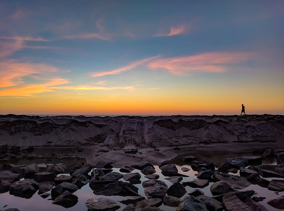 Silhouette Photo of Man Walking on Hill Near Body of Water and Rock Formations HD wallpaper