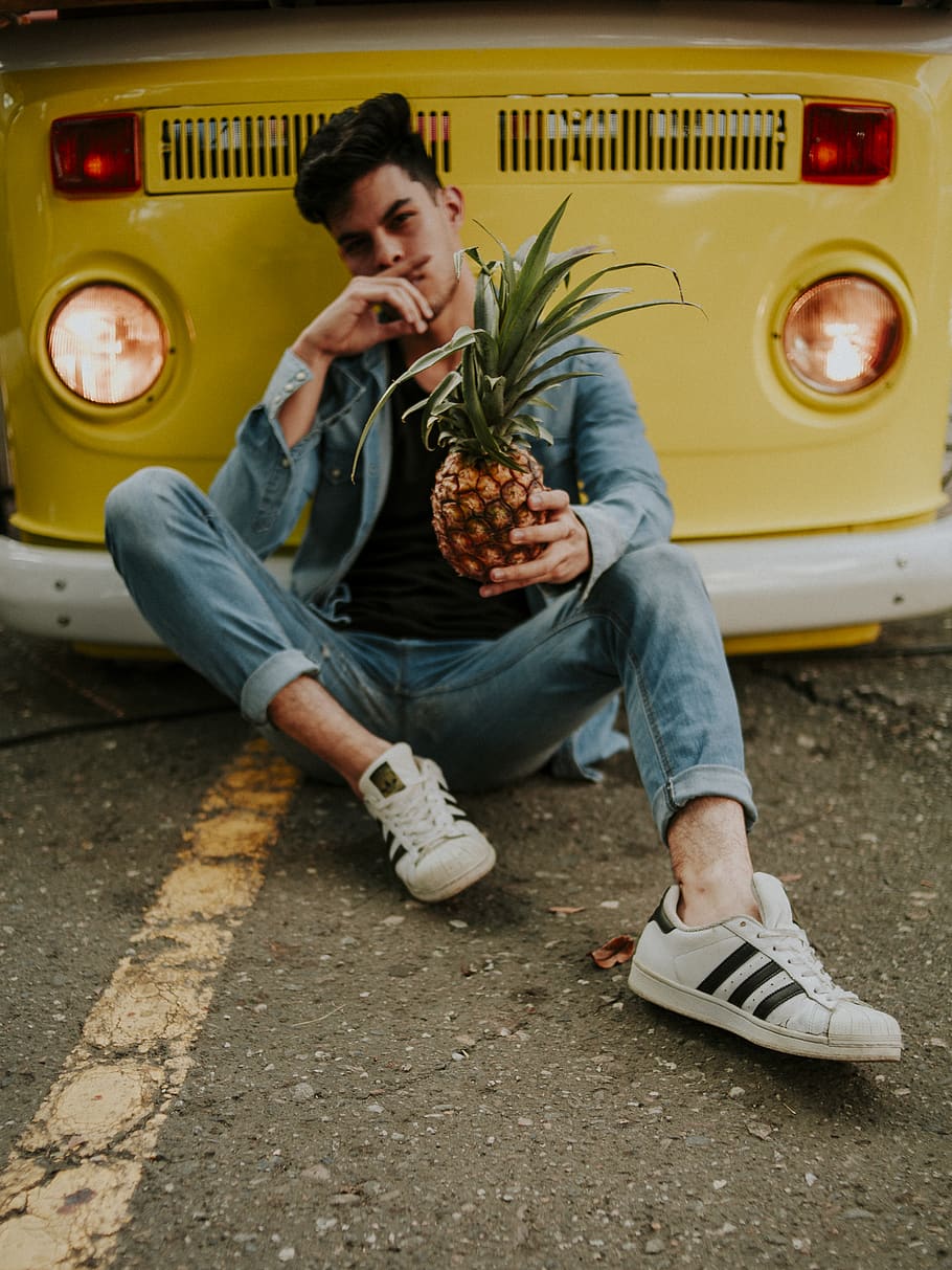 man holding pineapple while leaning on yellow van, footwear, apparel