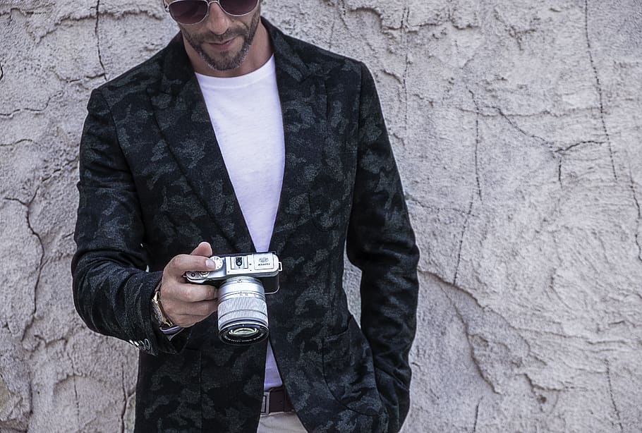 Man in Blue and Black Camouflage Coat Holding Silver Dslr Camera Standing in Front of Gray Concrete Wall