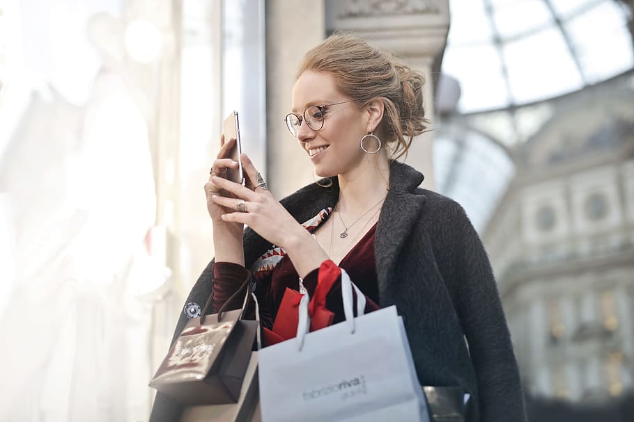 A young blond woman wearing a satin dress and an overcoat looks at her mobile phone while holding shopping bags in her hands
