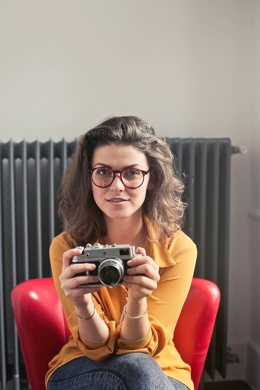 Young Woman Wearing Yellow Collared Shirt And Jeans With Spectacles Holding A Camera While Sitting at Red Chair