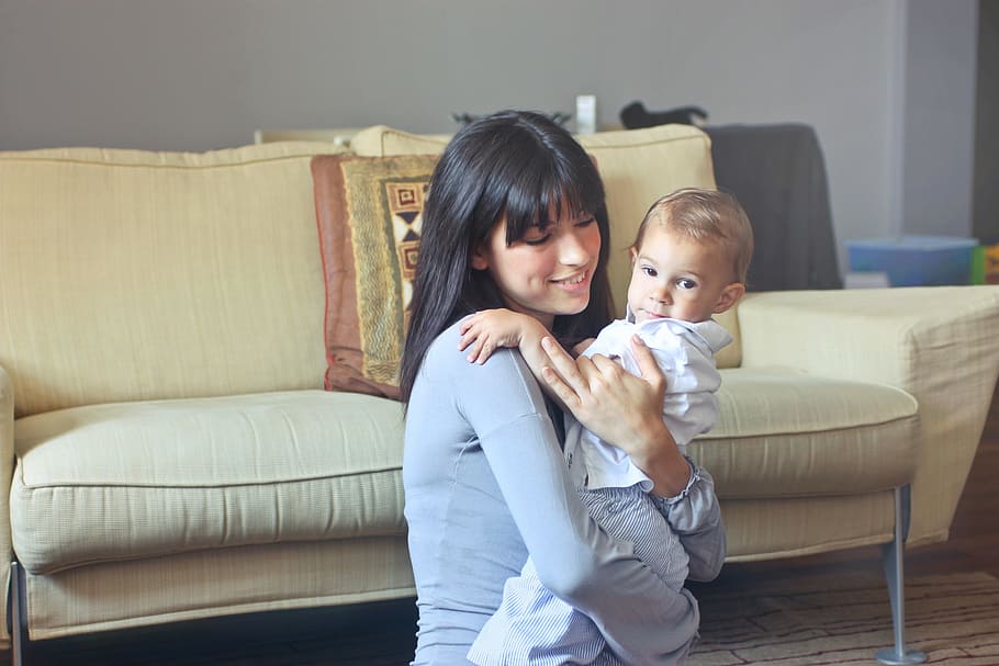 Beautiful young mother carrying her child in room with beige sofa in the background