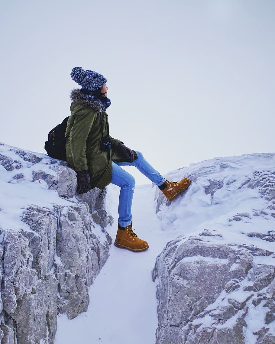 woman wearing hooded jacket, blue jeans, and brown boots sitting on rock covered with snow