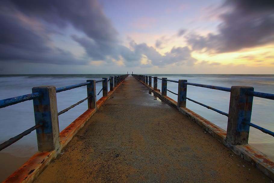 One Point Perspective Photo of Brown Concrete Bridge Leading to Body of Water With Gray Nimbus Clouds As Background during Golden Hour HD wallpaper