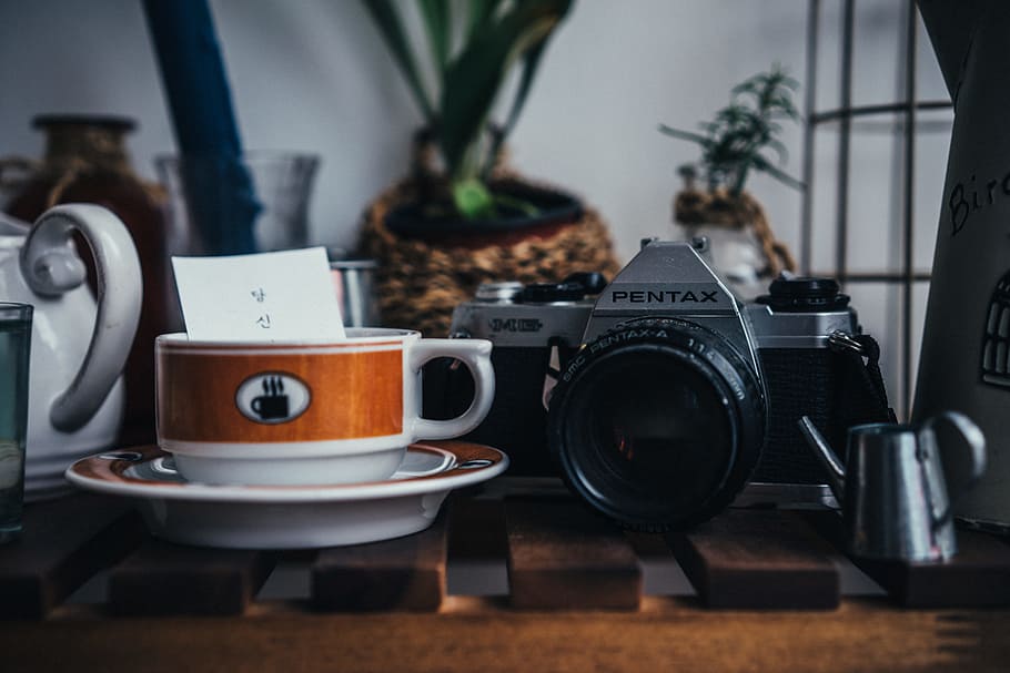 gray and black Pentax MILC camera beside round white and brown ceramic teacup on top of round white and brown saucer on top of table HD wallpaper