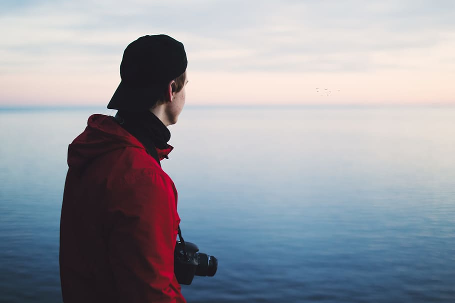 Man in Red Jacket Standing Near Body of Water, camera, cap, clouds HD wallpaper