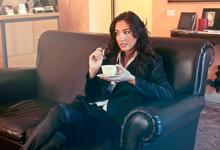 Woman in Black Blazer Holding Teacup and Spoon While Sitting on Two Seater Black Sofa