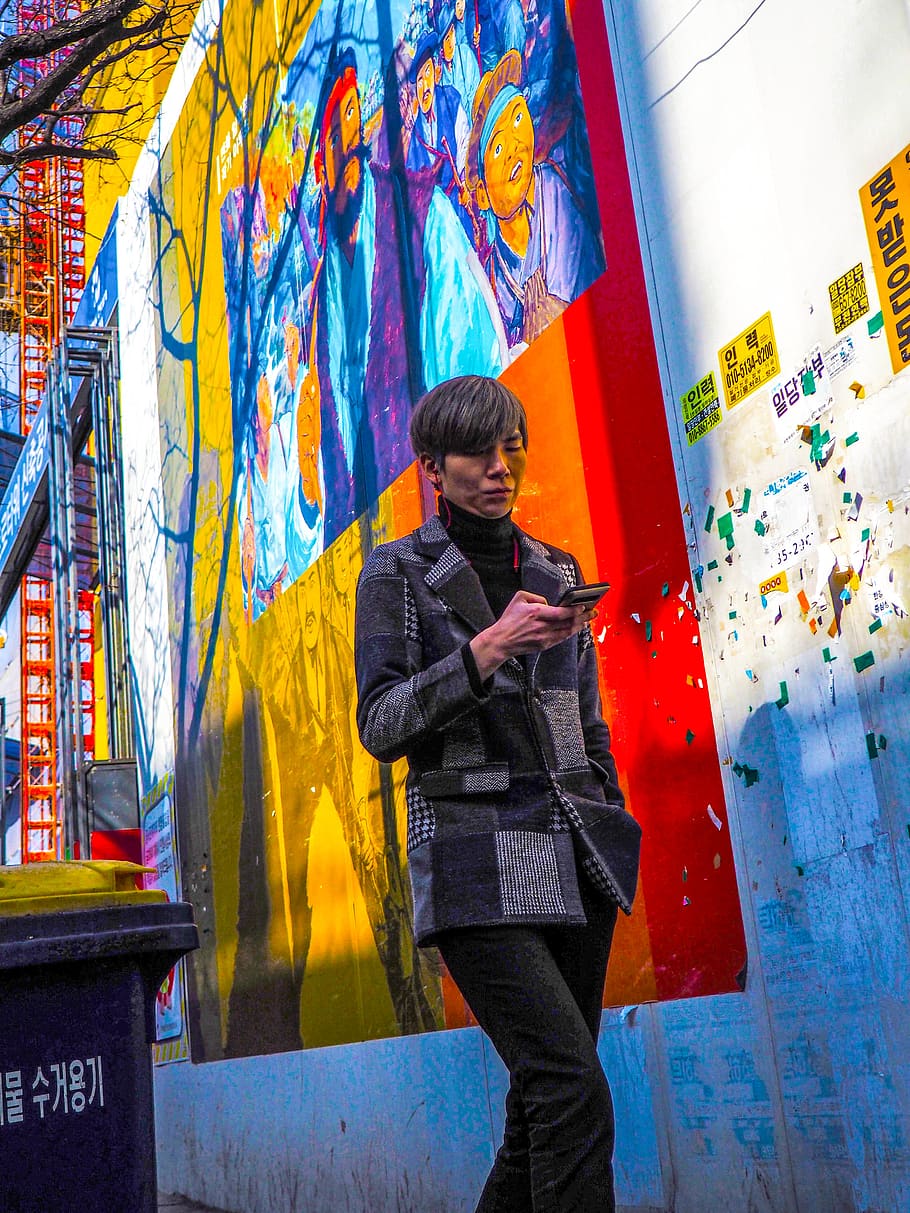 person in gray coat holding smartphone while walking near graffiti wall