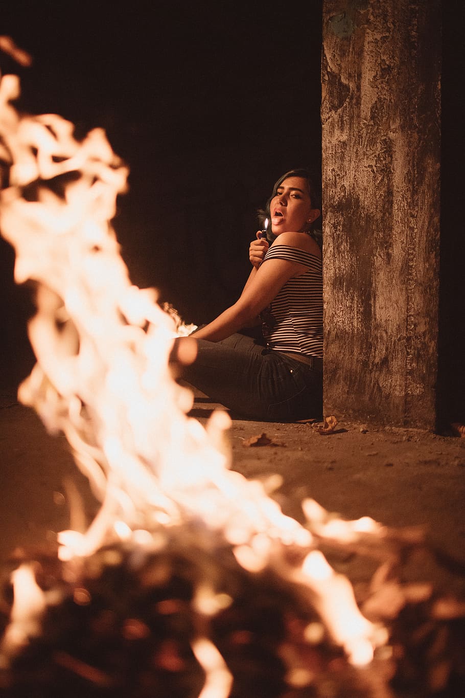 Woman Sitting on the Ground Beside Pillar Sticking Her Tongue Out Near Bonfire HD wallpaper