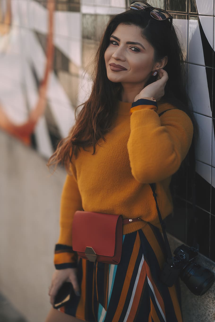 woman wearing mustard-colored sweater and striped skirt with hanged camera on shoulder leaning on wall