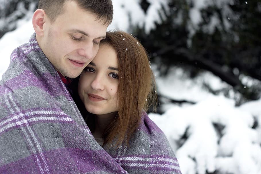 A happy man and woman wrapped in a blanket together next to a snow covered tree.