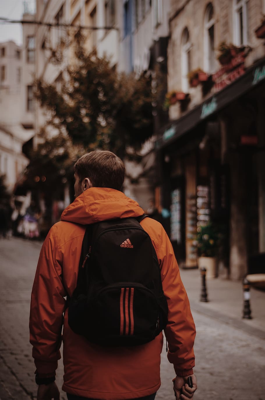 Man in Orange Jacket With Black Adidas Backpack Walks in Middle of Road