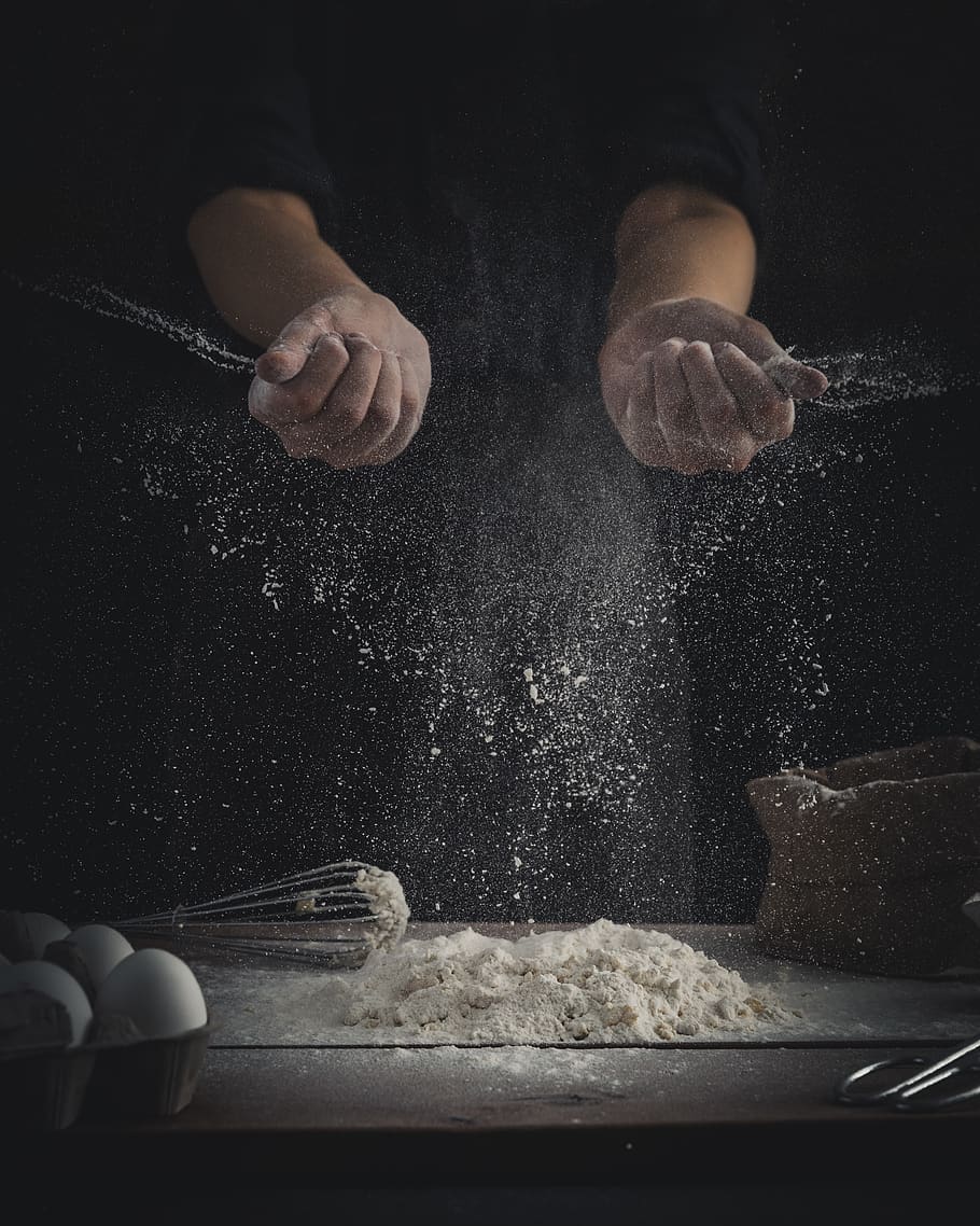 person pouring flour on table beside eggs and whisk, one person