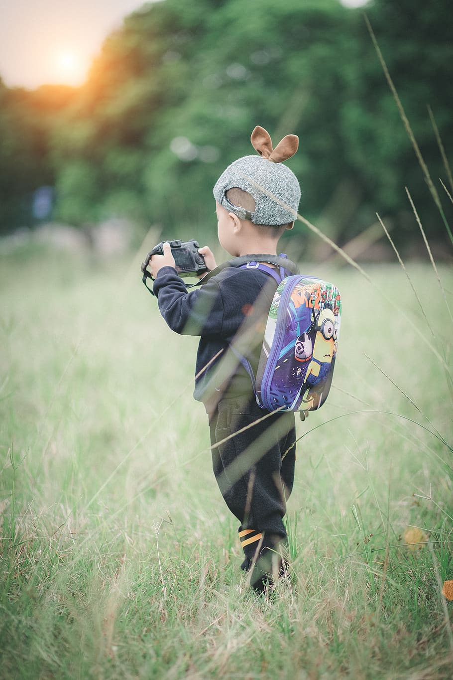 Boy Carrying Backpack Holding Camera Standing on Green Grass HD wallpaper